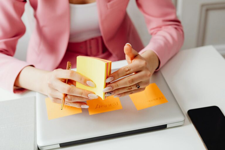 A woman in a pink blazer arranges sticky notes on her laptop, focusing on organization and planning.