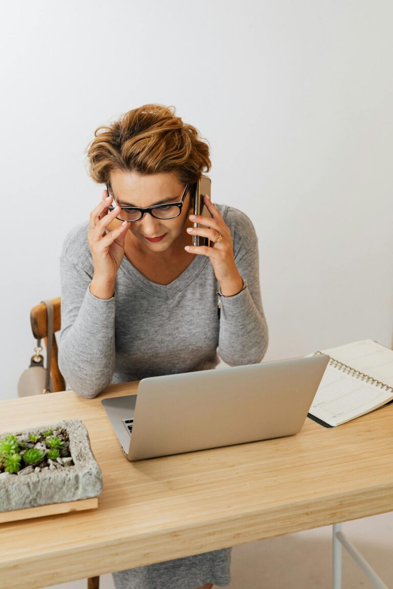 Woman multitasking between phone call and laptop at wooden desk in modern workspace.